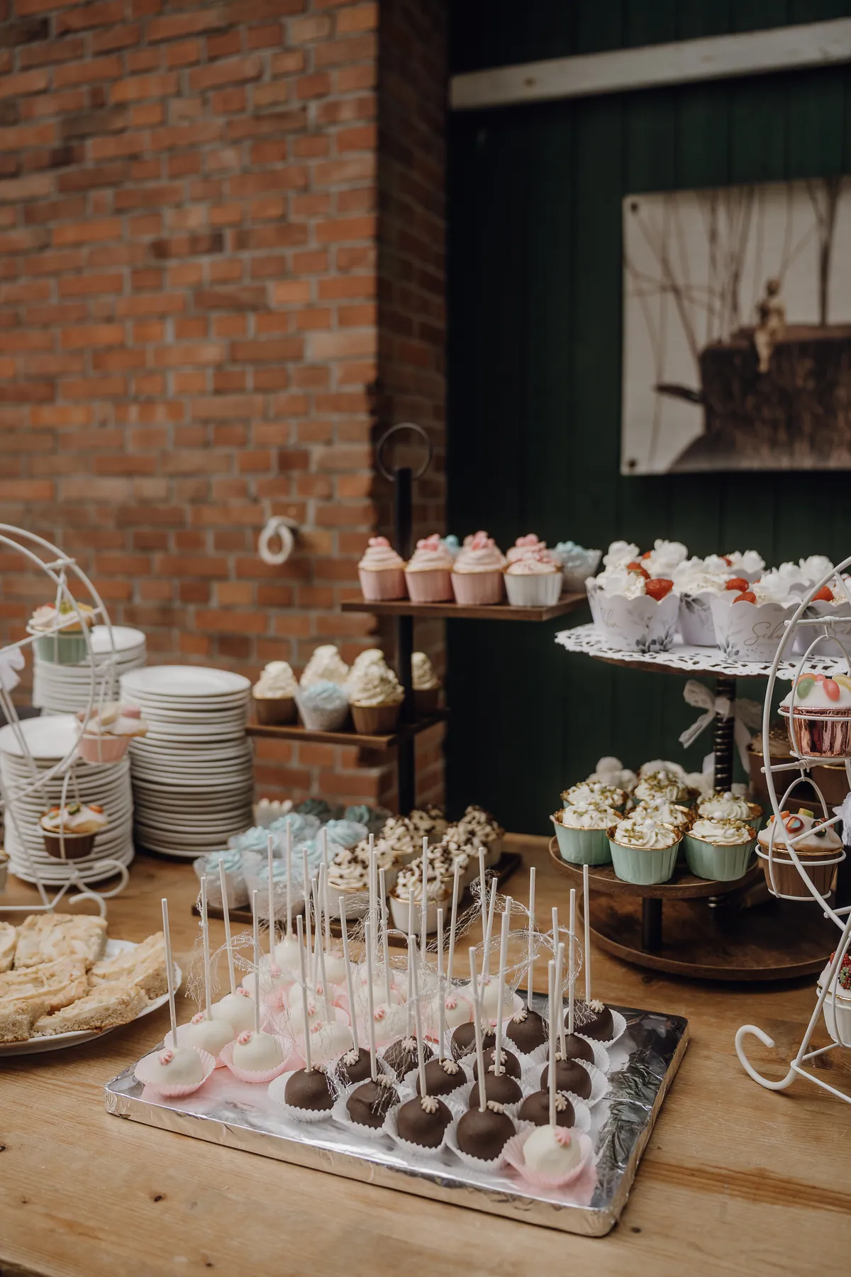 Das Kuchenbuffet: weiße und schokoladige Cake-Pops auf einem Silbertablett, Cupcakes auf Etageren und gestapelte Teller vor einer Backsteinwand.