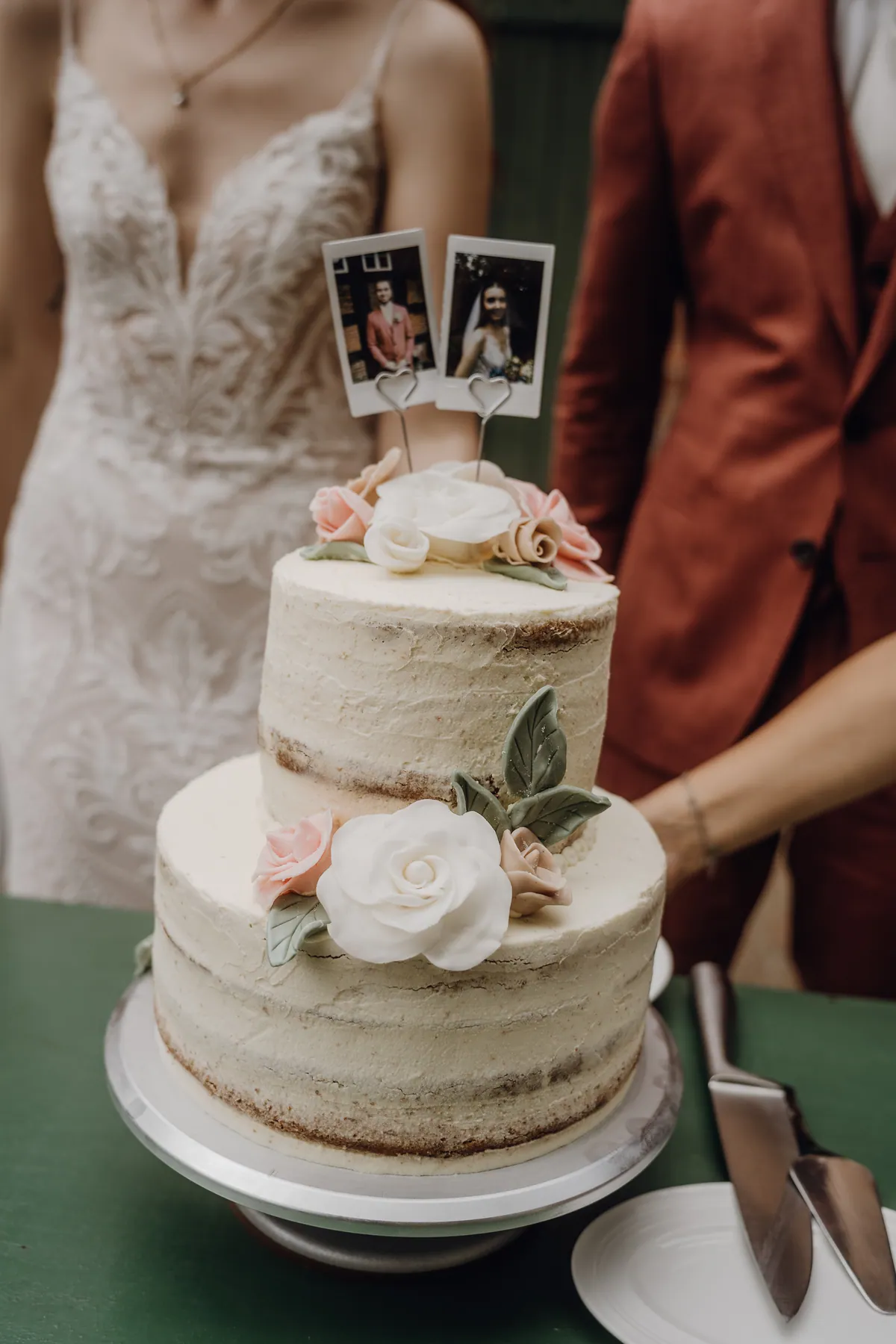 Die zweistöckige Hochzeitstorte mit Zuckerrosen trägt zwei Sofortbilder von Braut und Bräutigam als Topper, dahinter stehen die beiden angeschnitten.