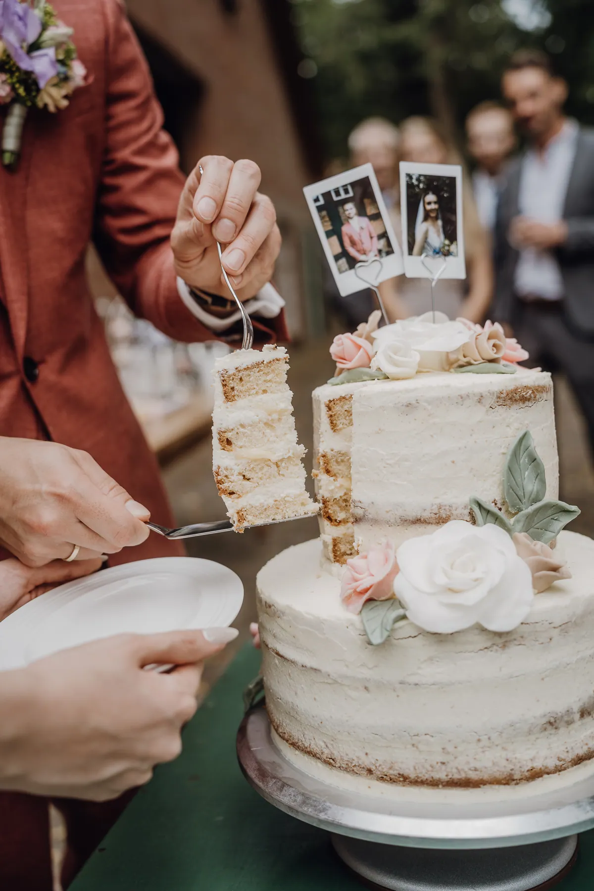 Ein Stück Torte mit hellen Biskuitschichten wird mit Tortenheber und Gabel herausgehoben, daneben die Sofortbild-Topper, im Hintergrund unscharf Gäste.
