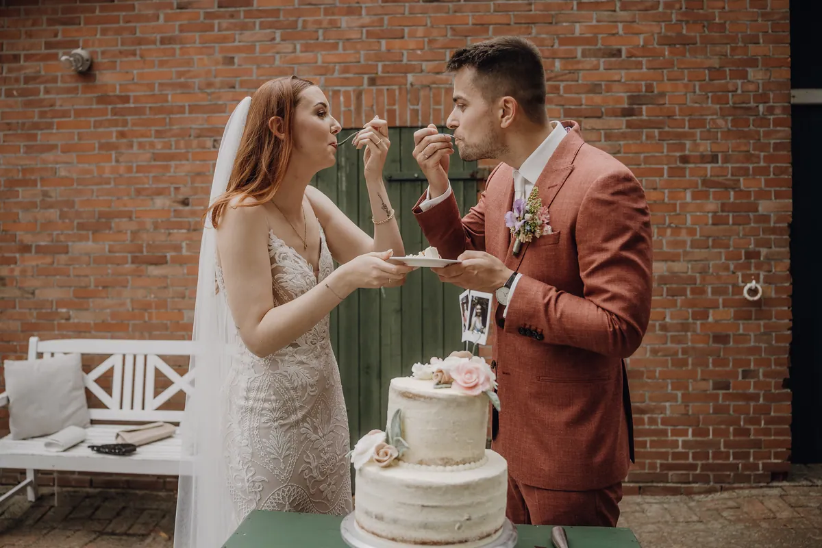 Theresa und Marvin stehen sich gegenüber und probieren gleichzeitig das erste Stück Torte von einem gemeinsamen Teller, hinter ihnen die Backsteinwand.