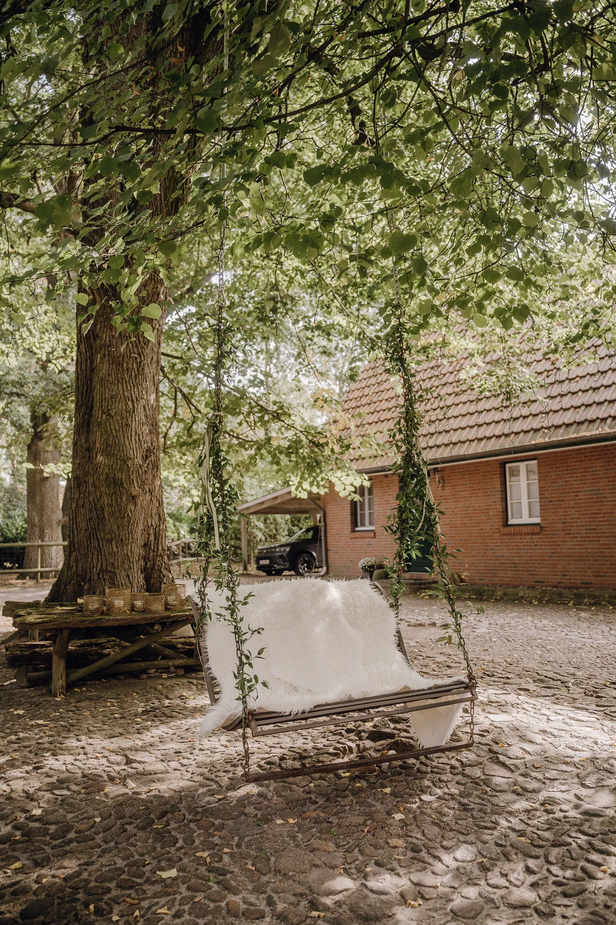 Eine Hollywoodschaukel mit weißem Fell hängt an begrünten Ketten unter einem großen Baum, dahinter ein Backsteinhaus und ein Hof aus Kopfsteinpflaster.