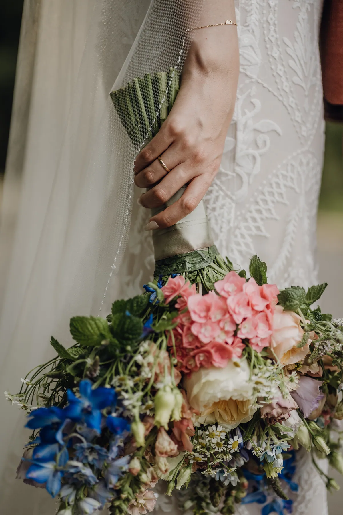 Nahaufnahme: Die Hand der Braut mit goldenem Ehering hält den Strauß aus blauem Rittersporn, rosa Blüten, Rosen, Kamille und Minze locker neben dem Spitzenkleid.
