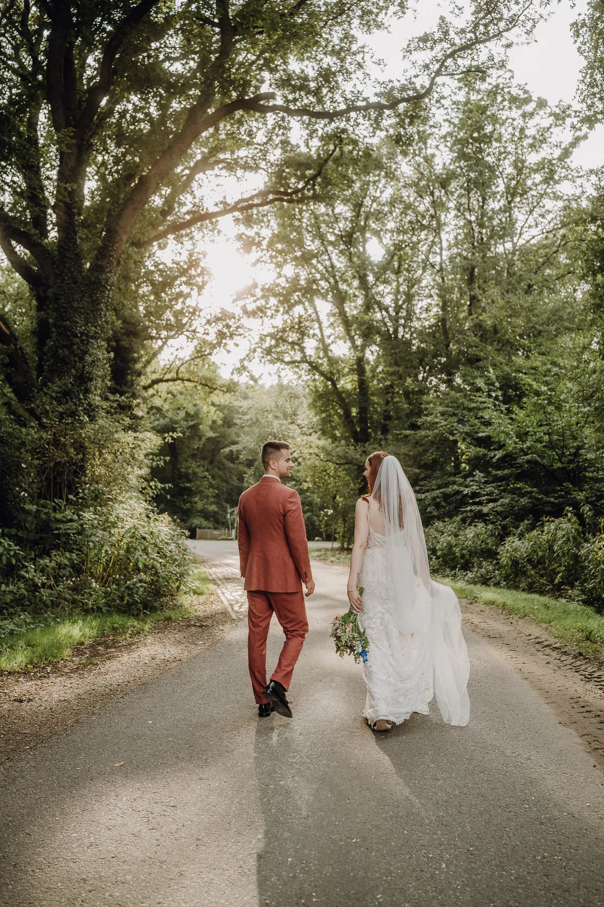 Das Paar geht auf einer Allee unter großen Bäumen davon und schaut sich dabei an, die Braut hält den Strauß, ihr Schleier weht im Gegenlicht.